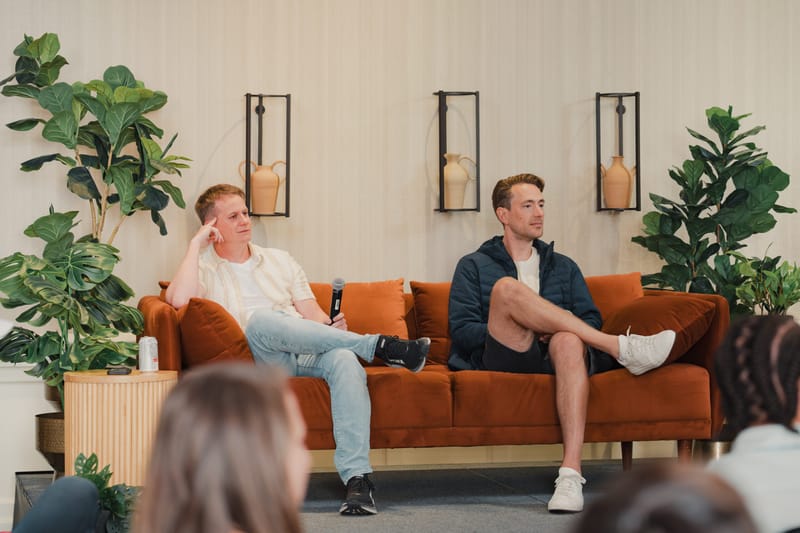 Two men in a fireside-chat on stage, seated on an orange sofa with one holding a microphone