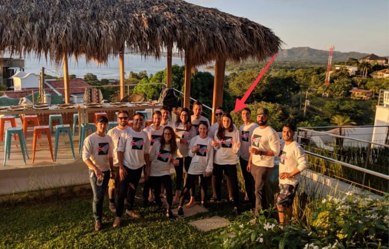 A team in matching shirts posing under a thatched palapa at sunset, with a red arrow pointing to one member