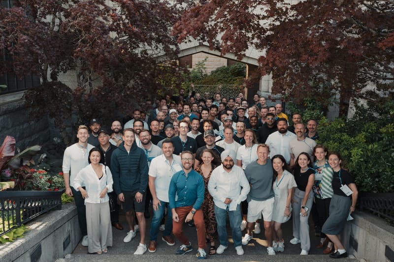 A large group of about fifty event attendees gathered for a photo on stone steps surrounded by greenery
